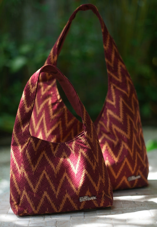 Two red and gold Batik-patterned bags on a wooden surface with a blurred green background.
