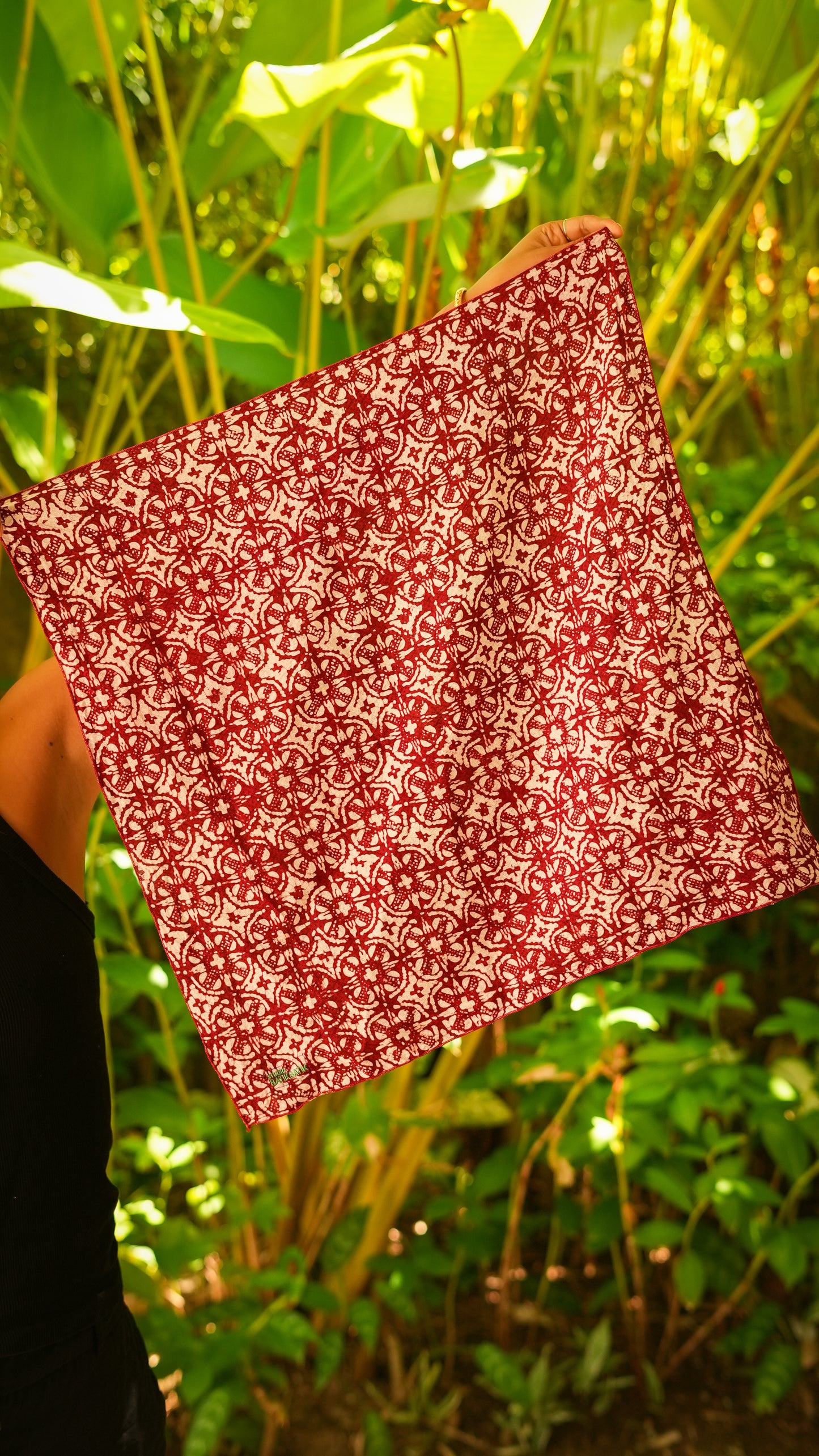 Red and white patterned batik bandana held in front of a green leafy background