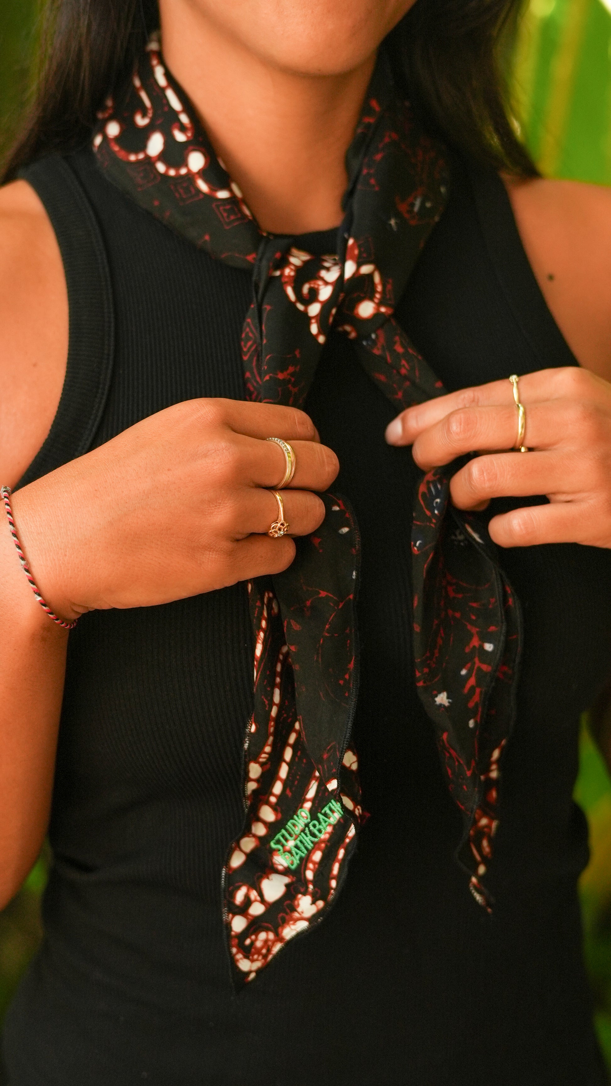 Person wearing a black sleeveless top with a batik patterned scarf, against a blurred green background with batik scarf