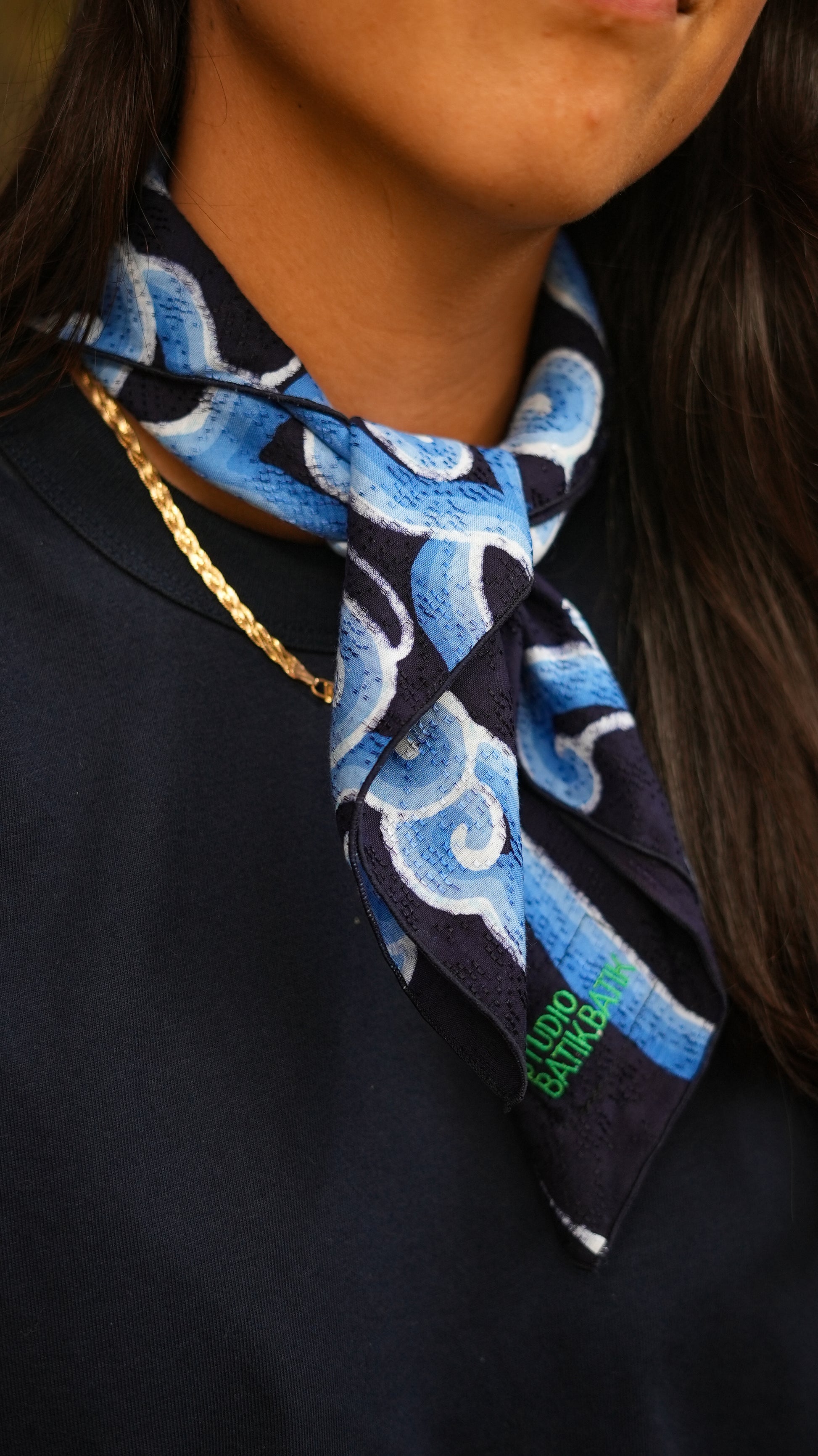 Person wearing a blue batik patterned scarf with a brand logo on a dark background