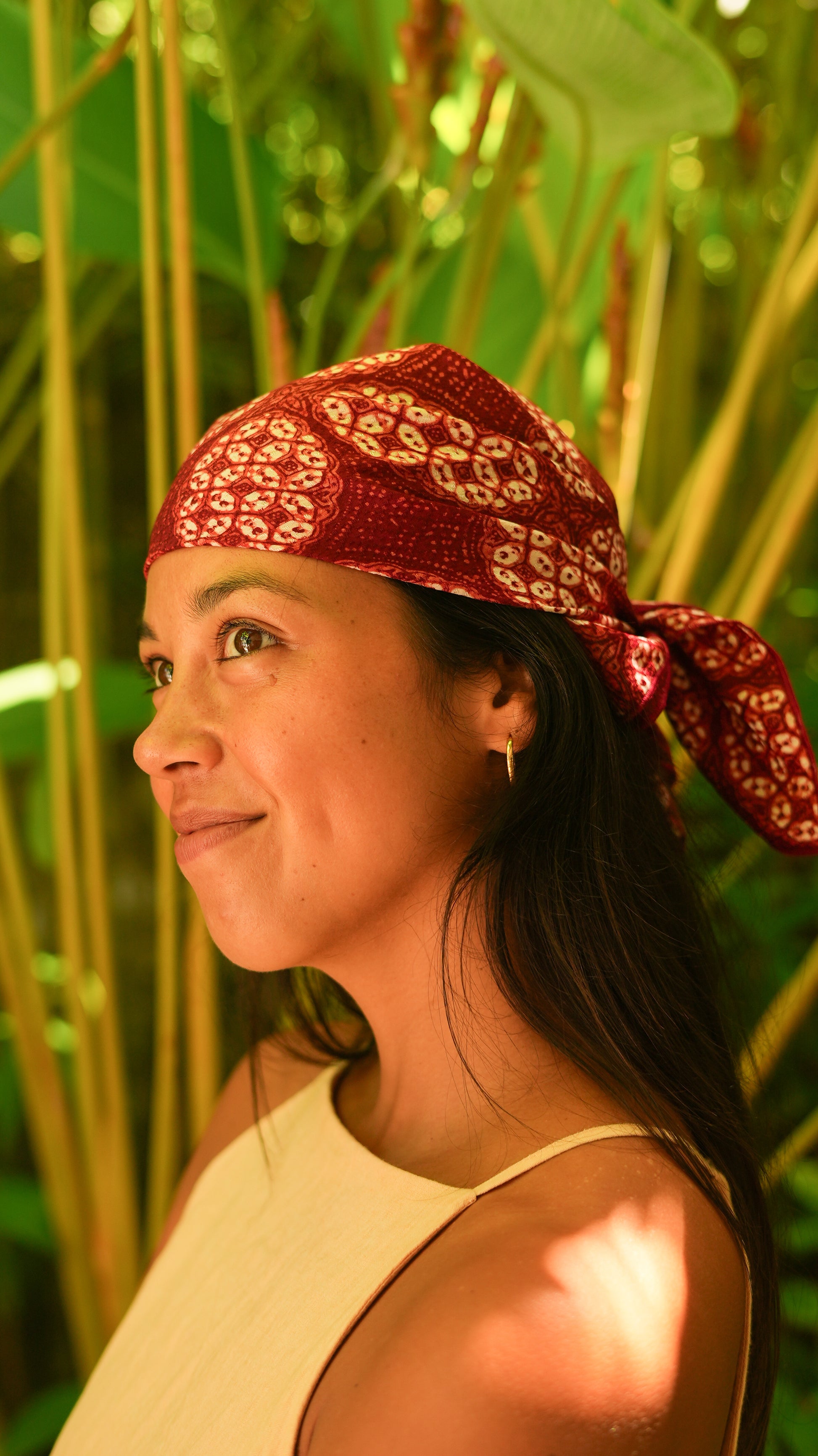 Woman wearing a red batik patterned headscarf in a natural setting with greenery.