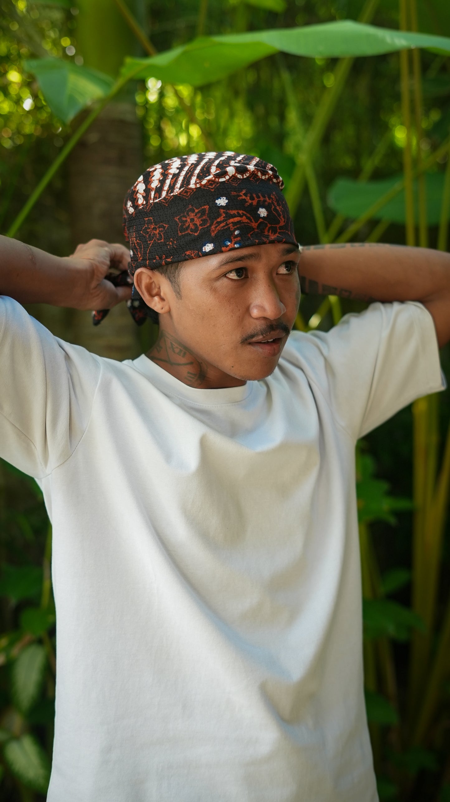 Man wearing a batik patterned parang headscarf in a natural setting with green foliage.