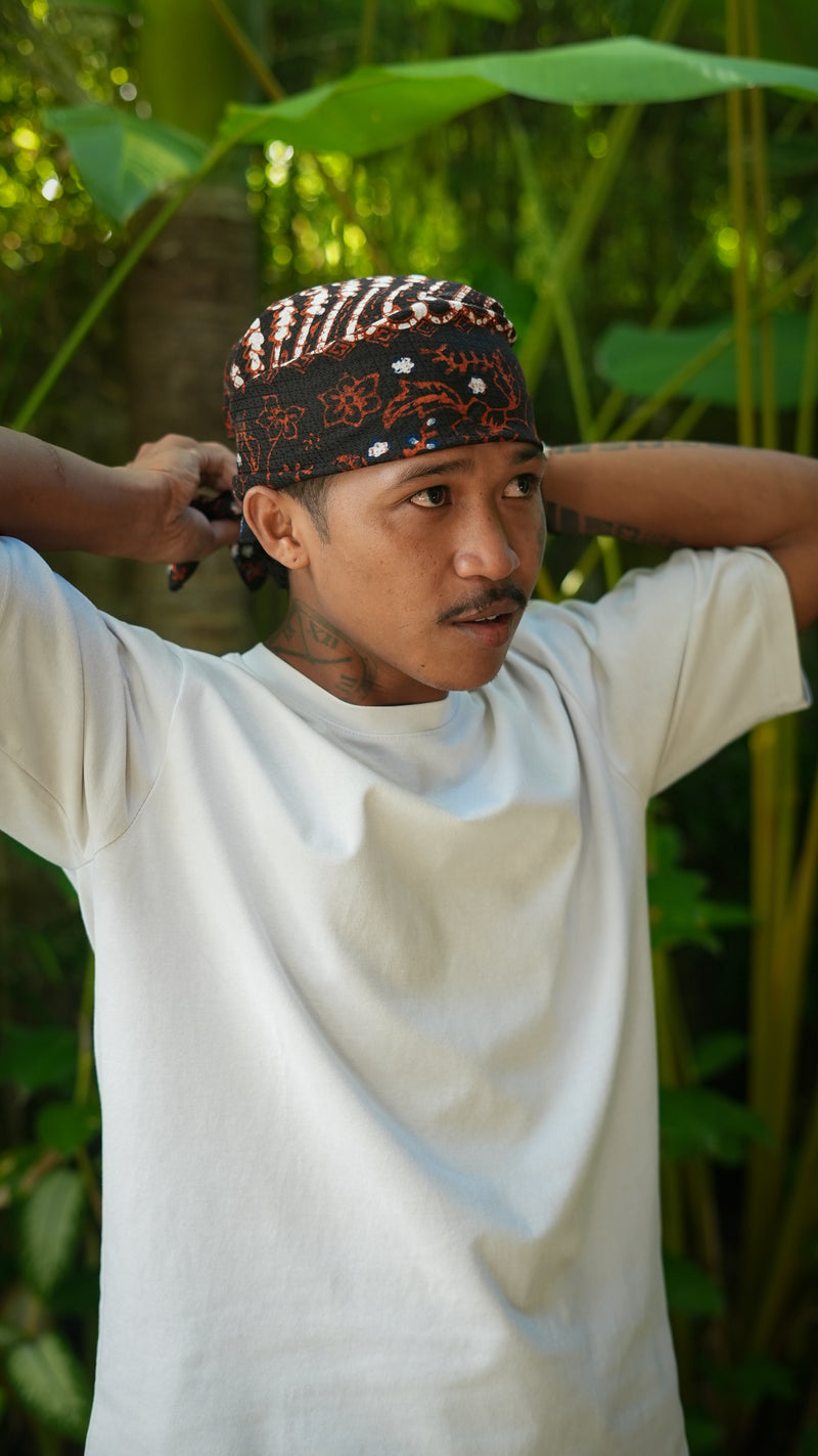 Man wearing a batik patterned parang headscarf in a natural setting with green foliage.