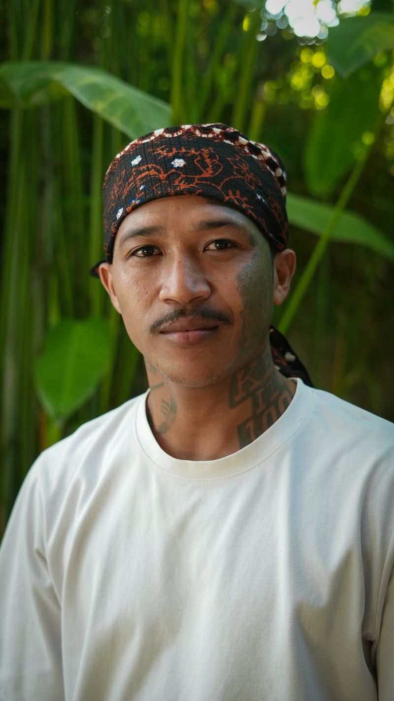 Man wearing a white t-shirt and batik parang patterned headscarf with green foliage in the background