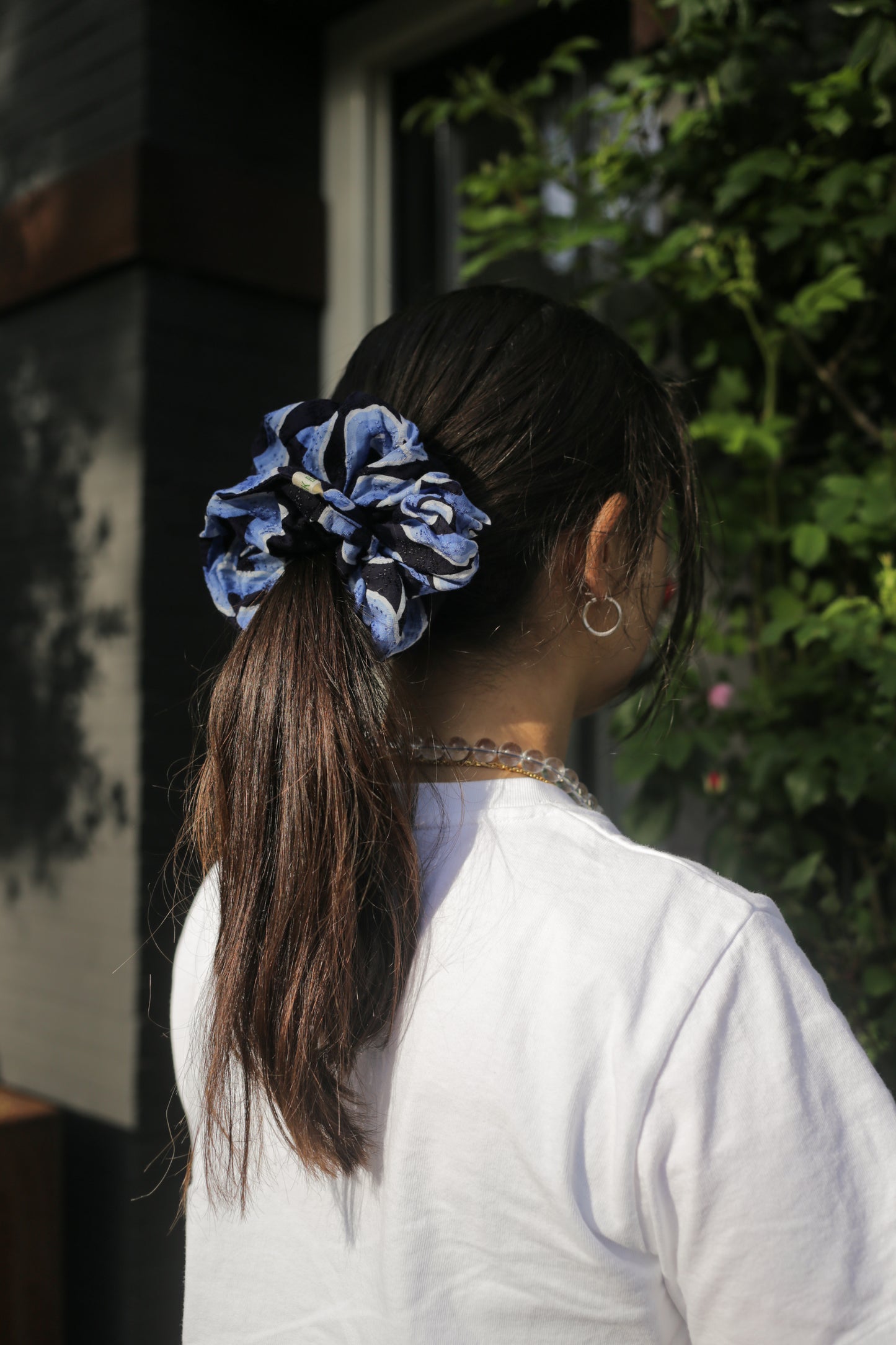 Person with a blue batik mega mendung scrunchie in their hair, standing outdoors with greenery in the background