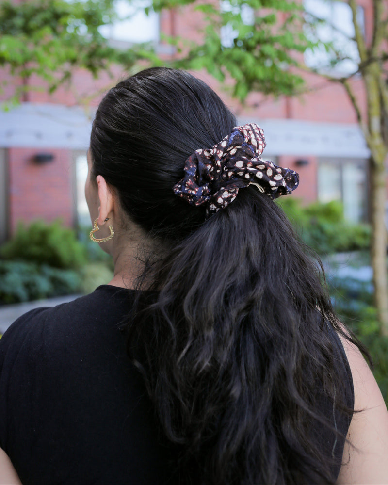 Person with a batik cap scrunchie in their hair, standing outdoors with greenery and a building in the background.