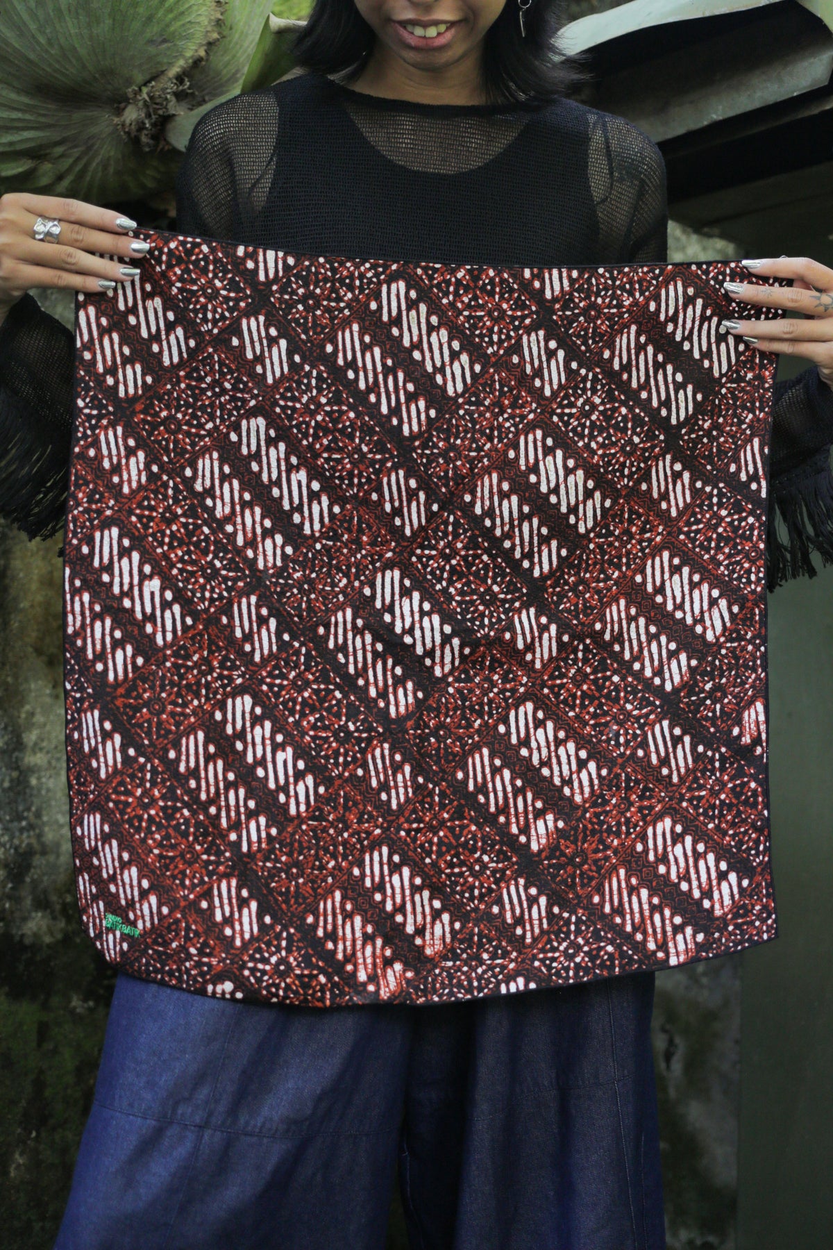 A woman holding up a square batik bandana panel with a red, black, and white geometric pattern, wearing a black sheer top and blue wide-leg pants.
