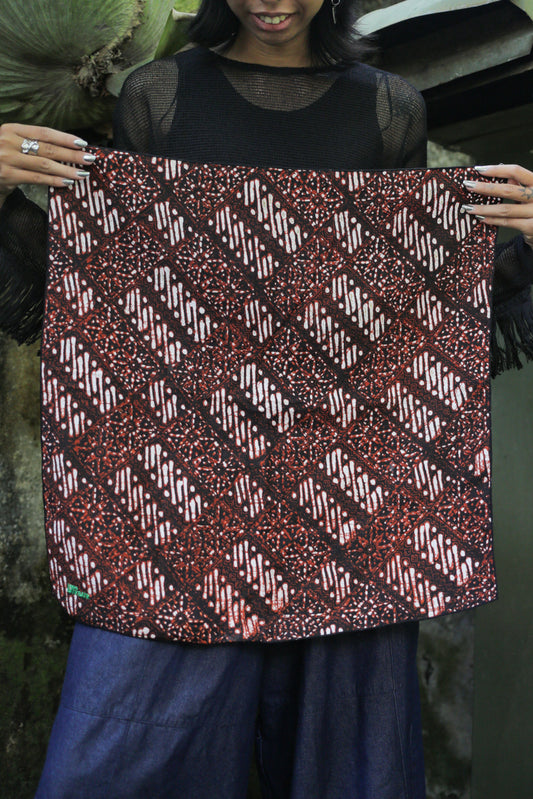 A woman holding up a square batik bandana panel with a red, black, and white geometric pattern, wearing a black sheer top and blue wide-leg pants.