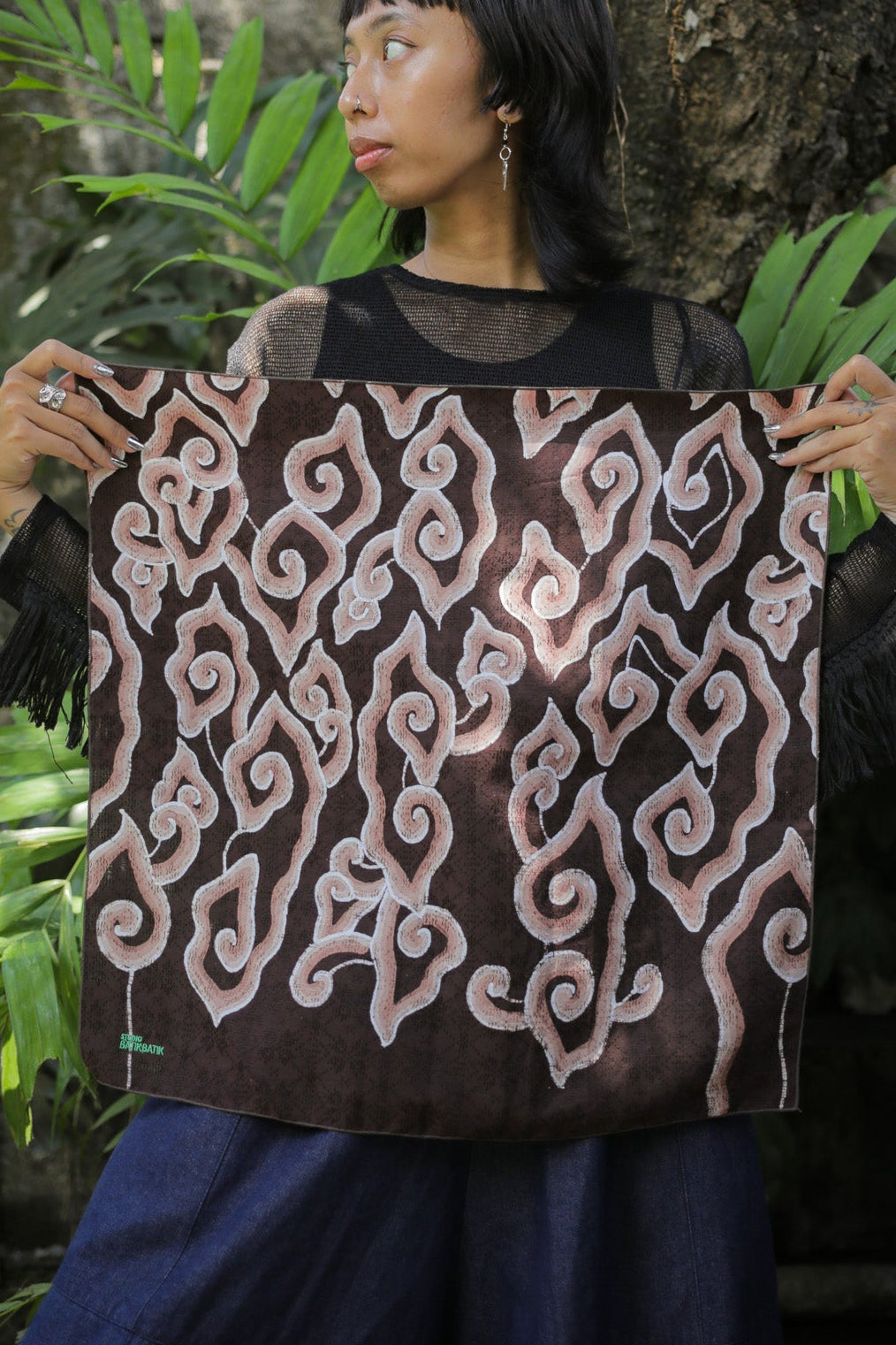 A woman stands outdoors in front of a tree and brick wall, holding up a square textile with a dark brown base and large light pink and white abstract scroll patterns.