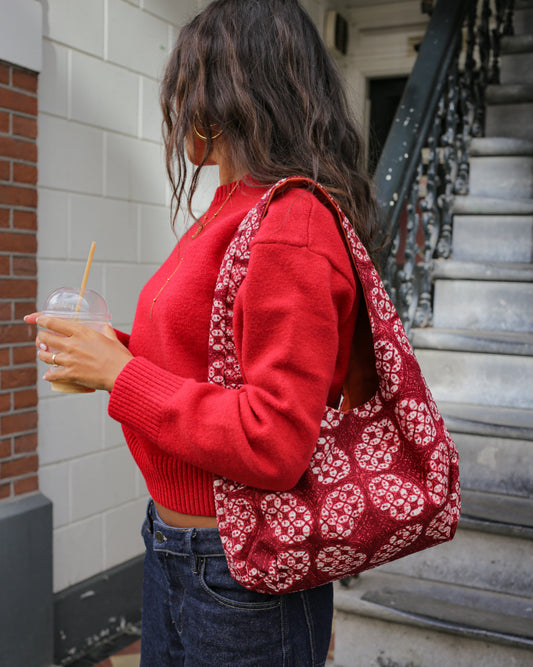 A woman wearing a red knit sweater and dark blue jeans carries a red and white handmade batik knit tote bag over her shoulder while standing on outdoor stone steps.