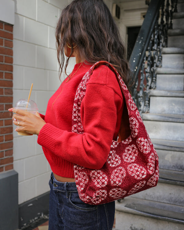 A woman wearing a red knit sweater and dark blue jeans carries a red and white handmade batik knit tote bag over her shoulder while standing on outdoor stone steps.